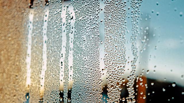 Close-up of a window with condensation, showing water droplets streaking down the glass. The blurred background reveals an outdoor scene with blue sky and faint structural or landscape elements.