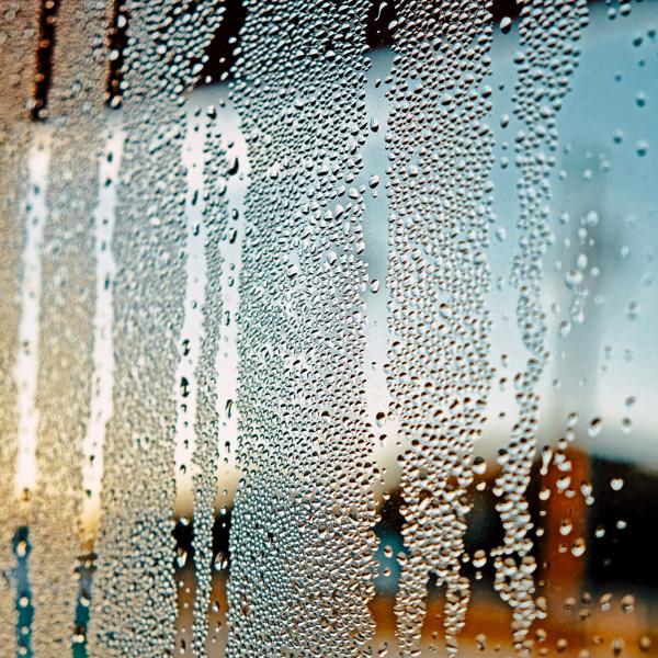 Close-up of a window with condensation, showing water droplets streaking down the glass. The blurred background reveals an outdoor scene with blue sky and faint structural or landscape elements.