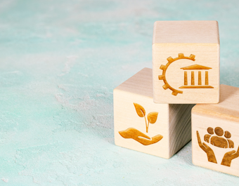 Three wooden blocks stacked on a light blue background, each engraved with a symbol: a gear and building for governance, hands holding a plant for sustainability, and hands cradling people for community support.