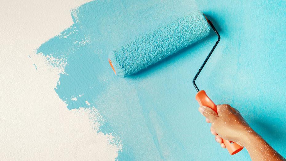 Hand using an orange-handled roller to apply light blue paint to a white wall.