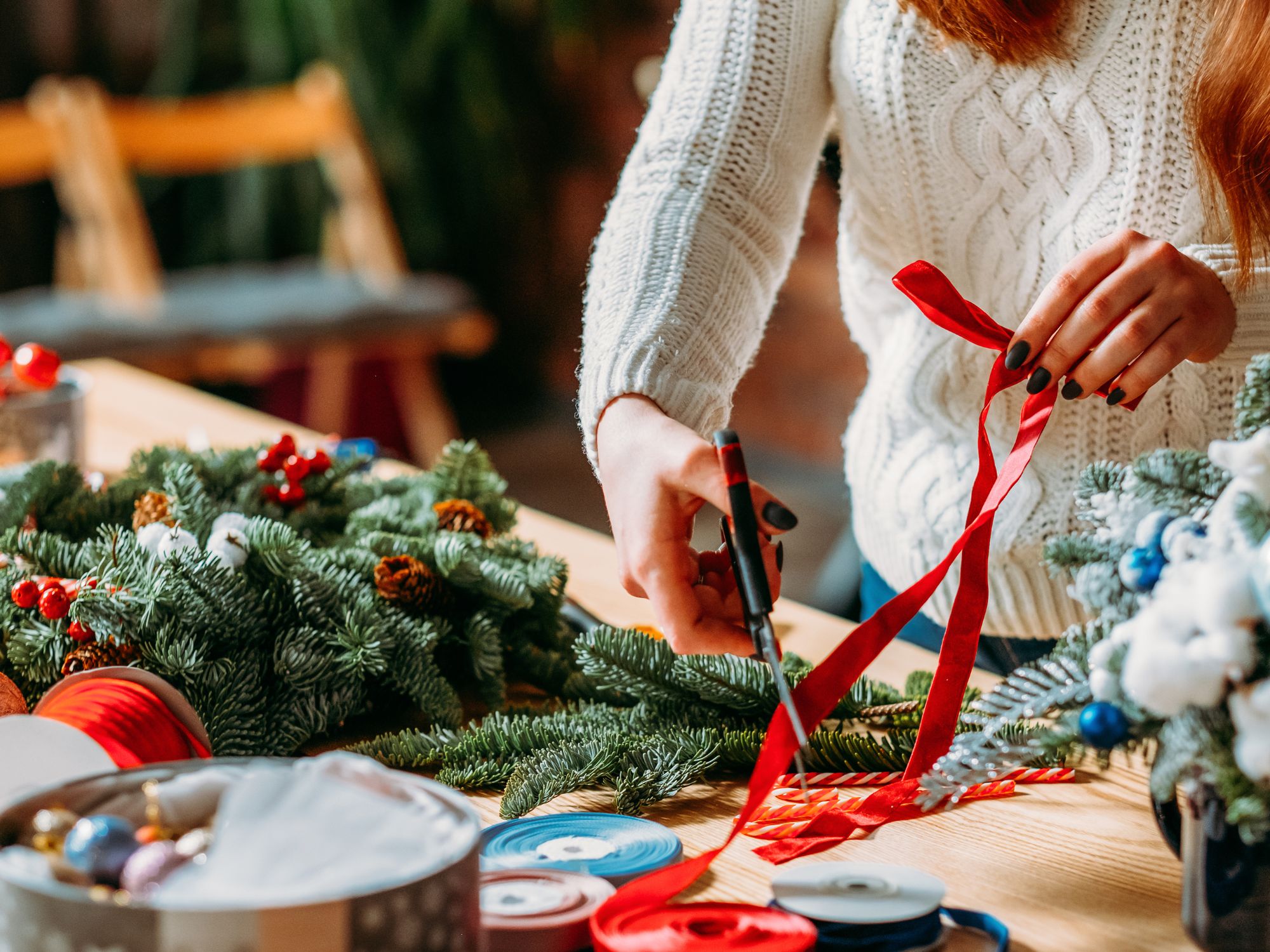 Close-up of someone crafting a Christmas wreath on a wooden table. The person is cutting a red ribbon with scissors, surrounded by evergreen branches, pinecones, and festive decorations like red and blue ribbons and ornaments.