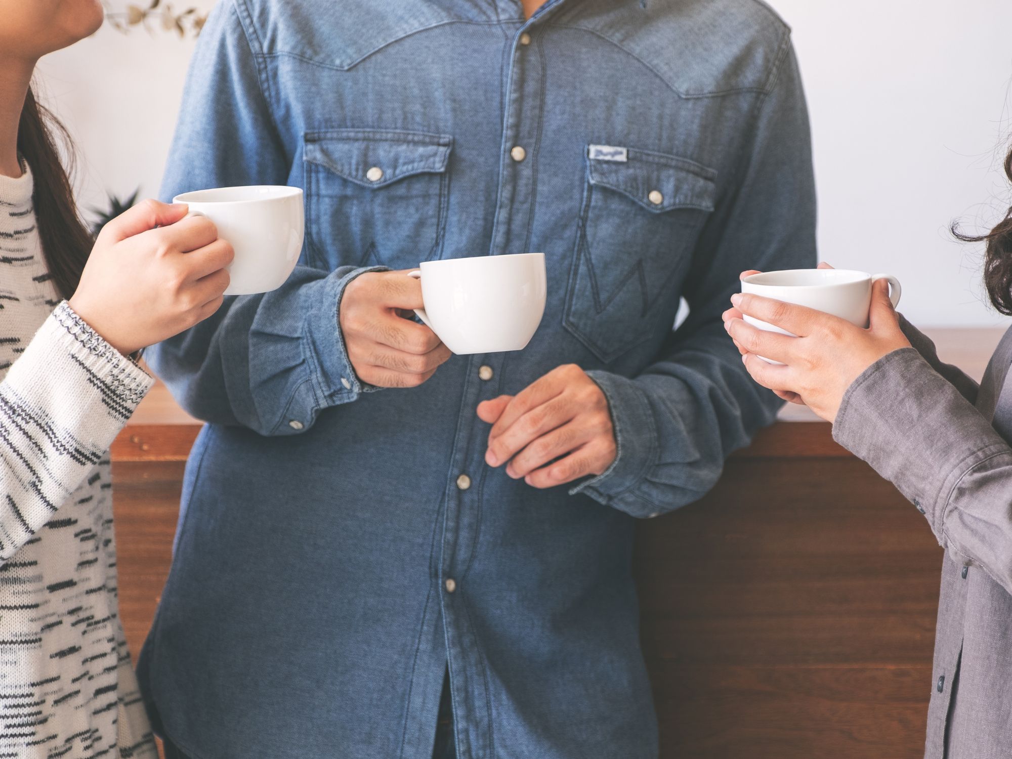Three people standing close together indoors, each holding a white ceramic mug. The person in the center is wearing a denim button-up shirt, while the others wear patterned and plain long-sleeve tops. They appear to be engaged in conversation, with a wooden counter and light-colored wall in the background.