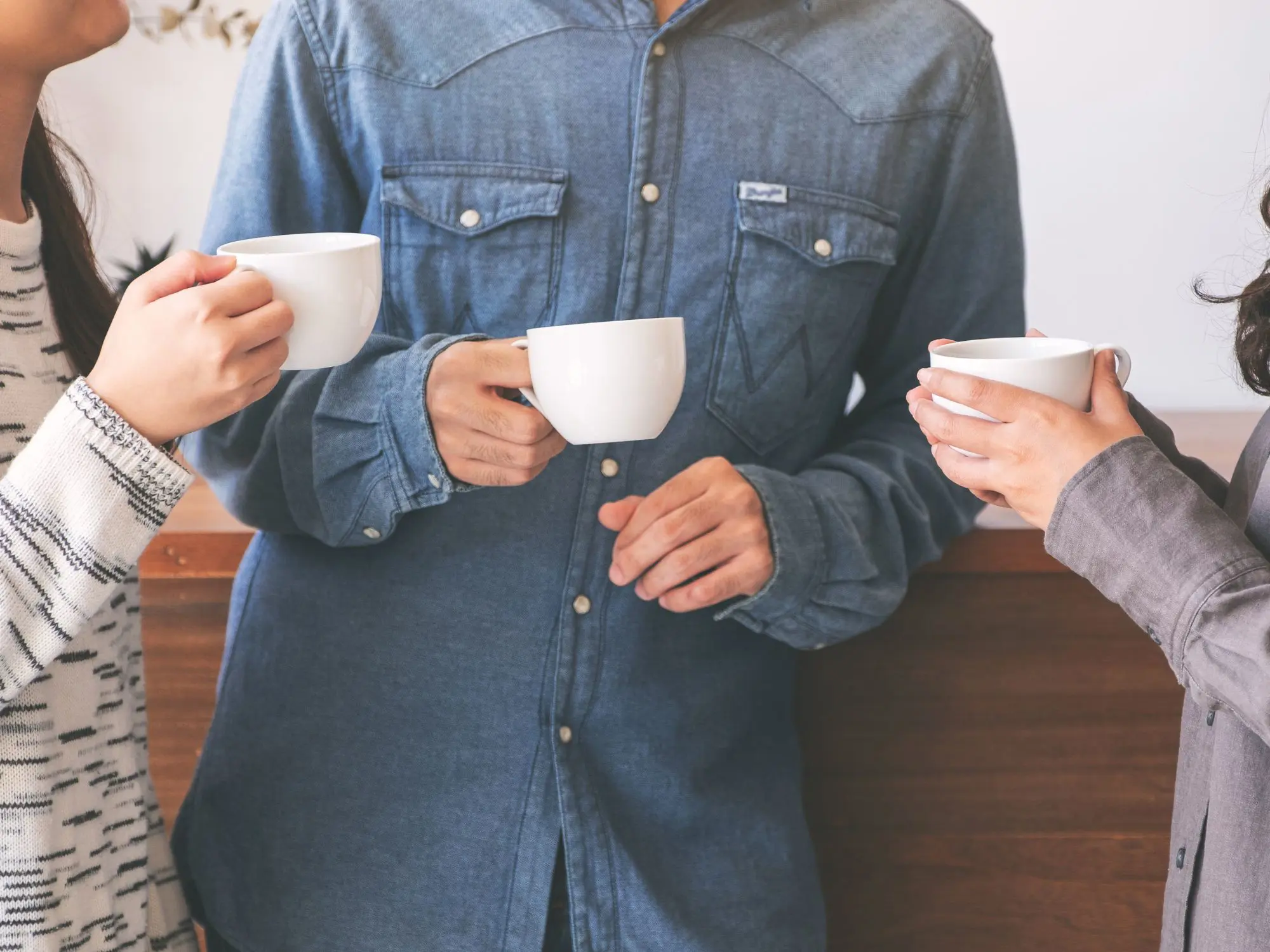 Three people standing close together indoors, each holding a white ceramic mug. The person in the center is wearing a denim button-up shirt, while the others wear patterned and plain long-sleeve tops. They appear to be engaged in conversation, with a wooden counter and light-colored wall in the background.