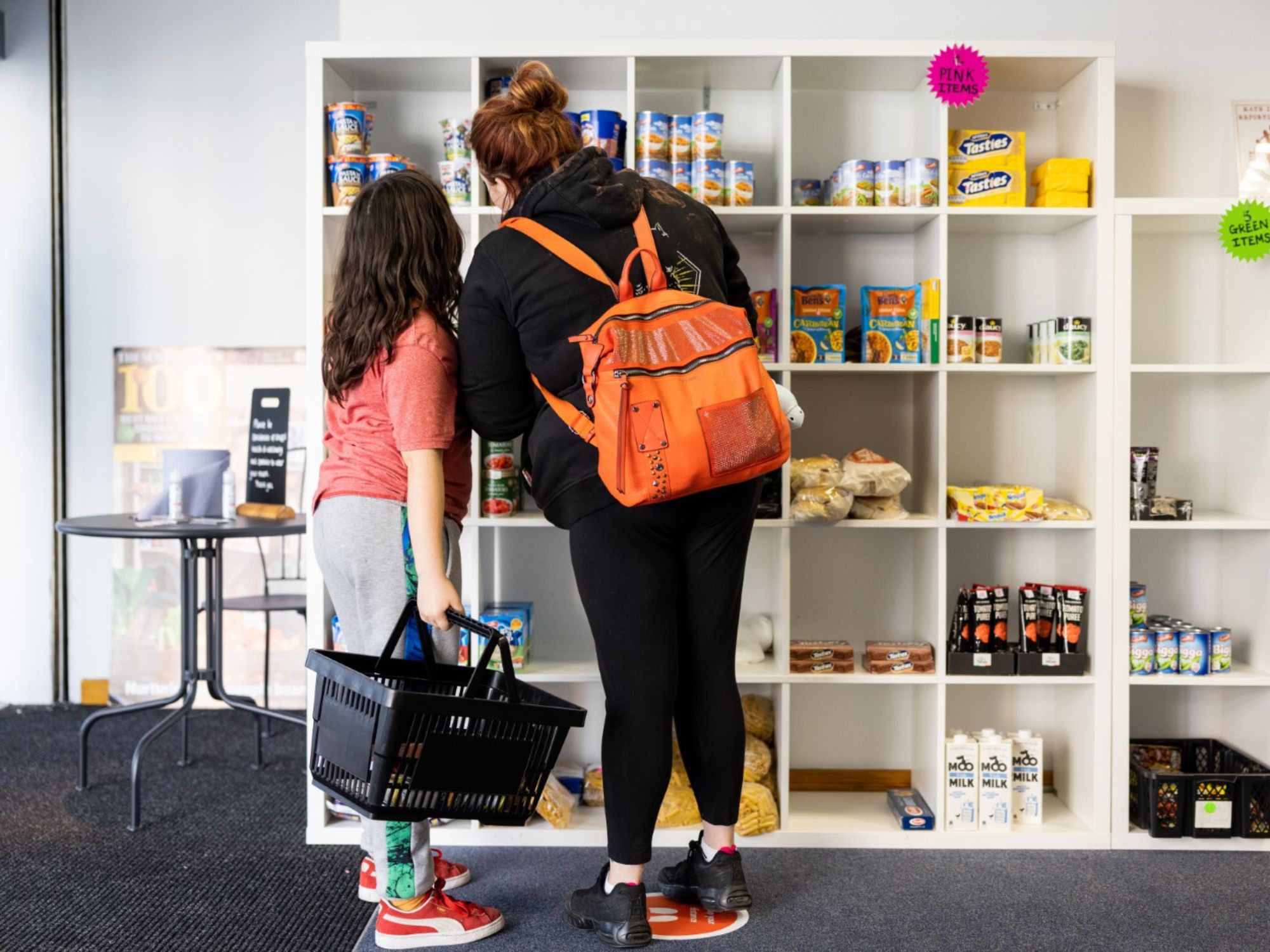Two people browsing shelves stocked with canned goods and boxed groceries at The Market Place; one holds a shopping basket, the other wears an orange backpack.