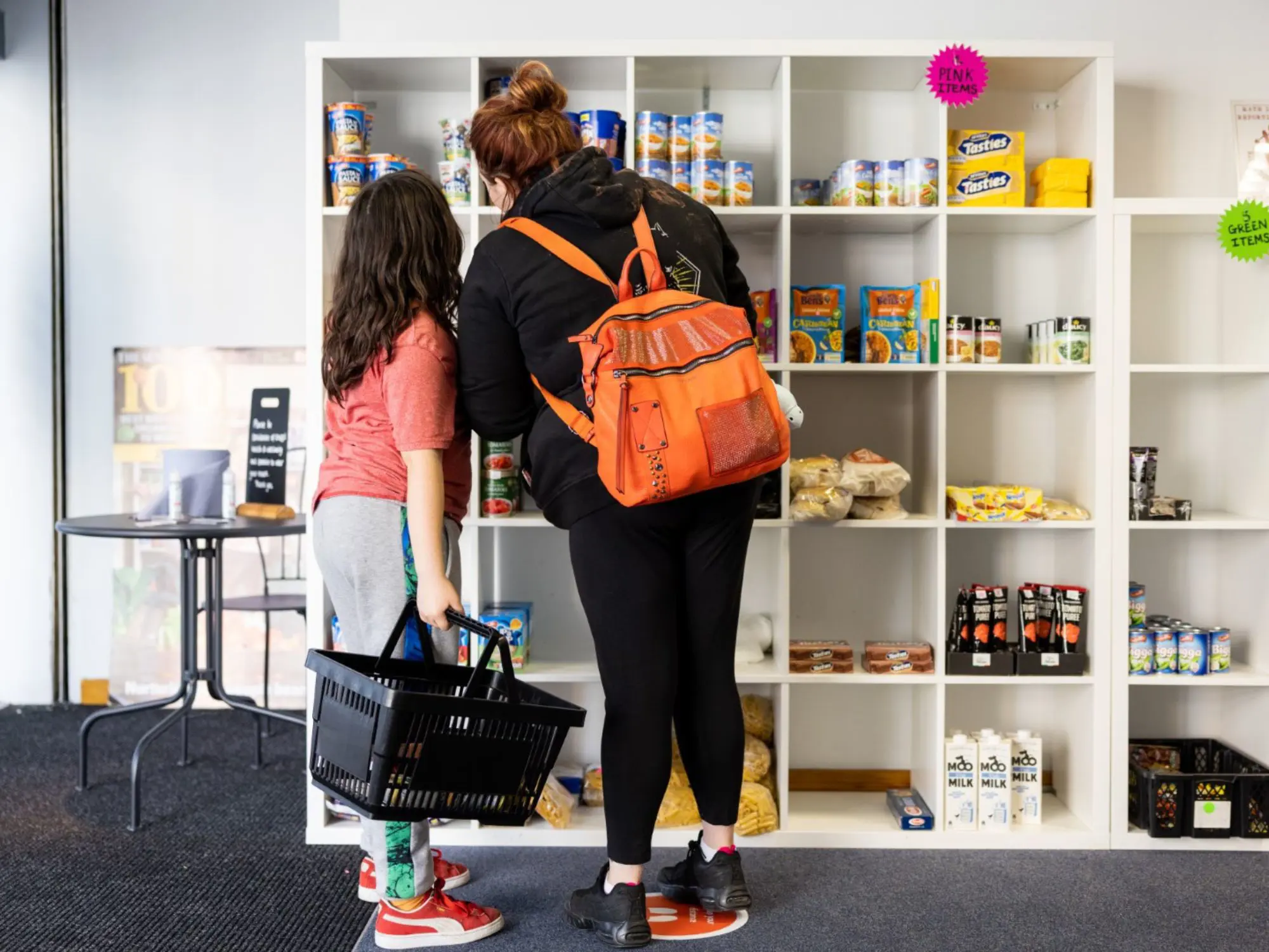 Two people browsing shelves stocked with canned goods and boxed groceries at The Market Place; one holds a shopping basket, the other wears an orange backpack.