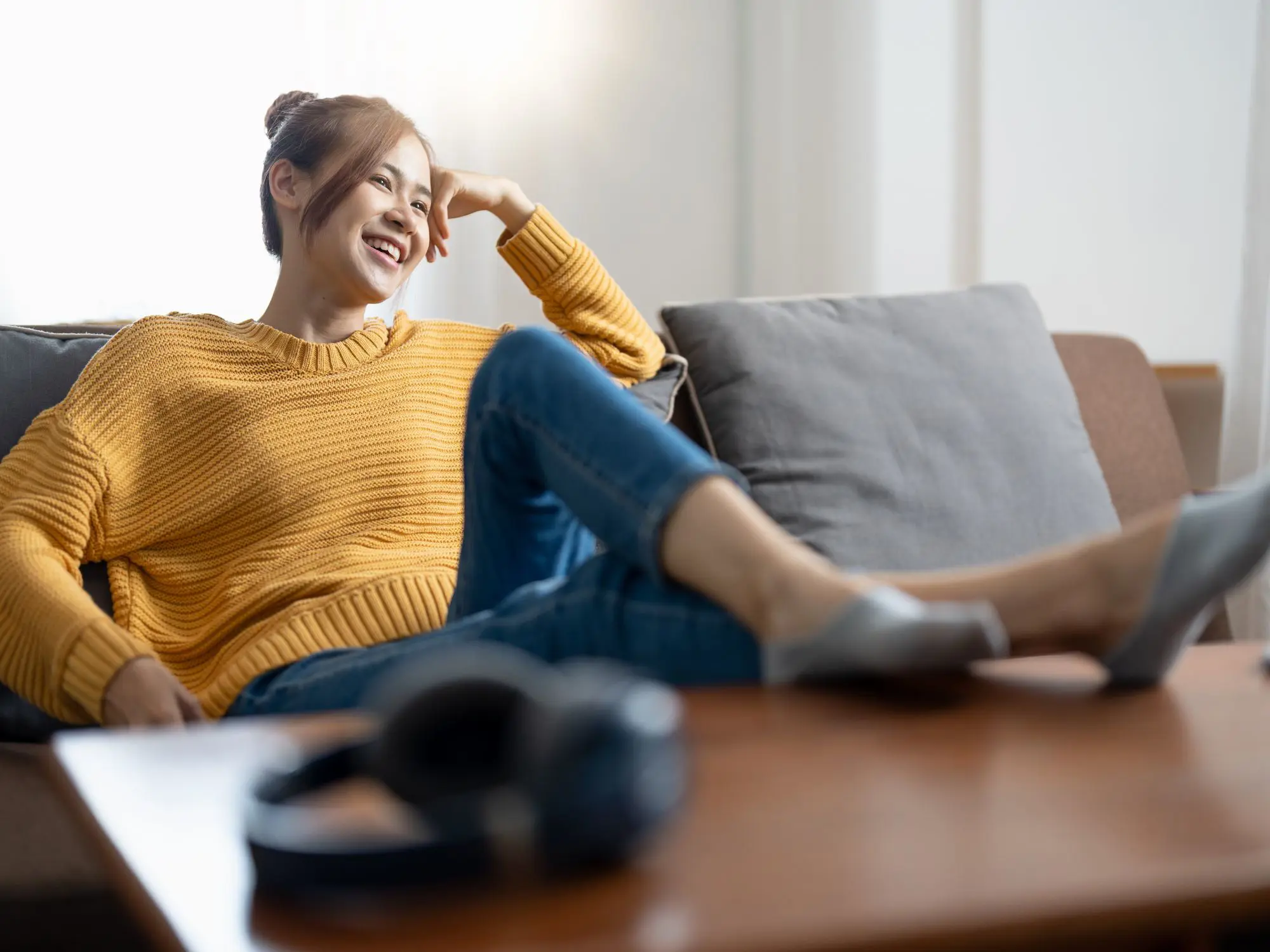 A person in a yellow sweater, blue jeans, and gray shoes sits on a couch with legs stretched out and crossed at the ankles; a pair of headphones rests on a table in the foreground.