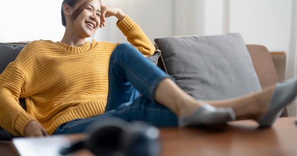 A person in a yellow sweater, blue jeans, and gray shoes sits on a couch with legs stretched out and crossed at the ankles; a pair of headphones rests on a table in the foreground.