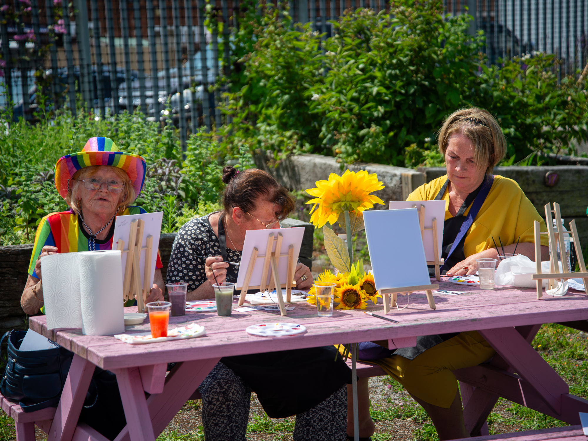 Three people sitting at a wooden picnic table outdoors, painting small canvases on easels surrounded by greenery, with paint pots, brushes and a sunflower decoration on the table.