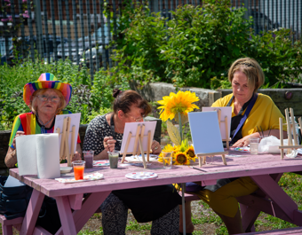 Three people sitting at a wooden picnic table outdoors, painting small canvases on easels surrounded by greenery, with paint pots, brushes and a sunflower decoration on the table.