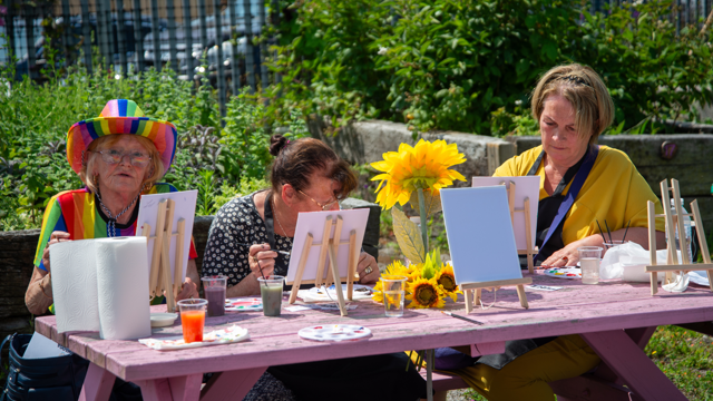 Three people sitting at a wooden picnic table outdoors, painting small canvases on easels surrounded by greenery, with paint pots, brushes and a sunflower decoration on the table.