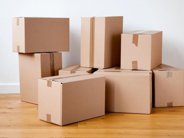 Eight sealed cardboard boxes of varying sizes stacked on a wooden floor against a plain white wall. Eight sealed cardboard boxes of varying sizes stacked on a wooden floor against a plain white wall.