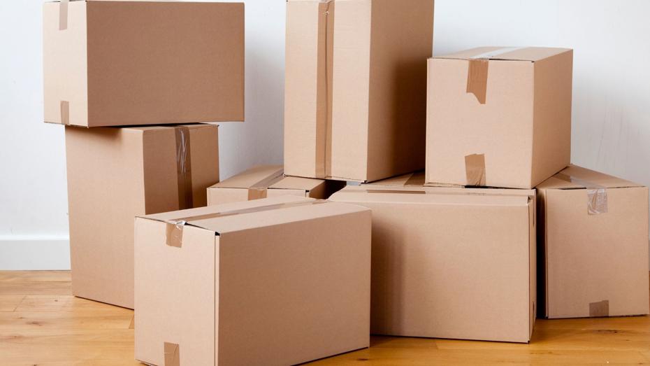 Eight sealed cardboard boxes of varying sizes stacked on a wooden floor against a plain white wall. Eight sealed cardboard boxes of varying sizes stacked on a wooden floor against a plain white wall.