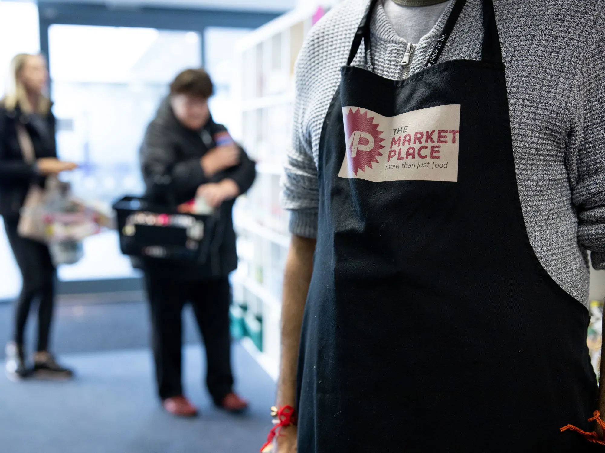 Person wearing an apron with ‘THE MARKET PLACE – more than just food’ logo, standing in a store with shelves and products; two other people are visible in the background, one holding a basket.