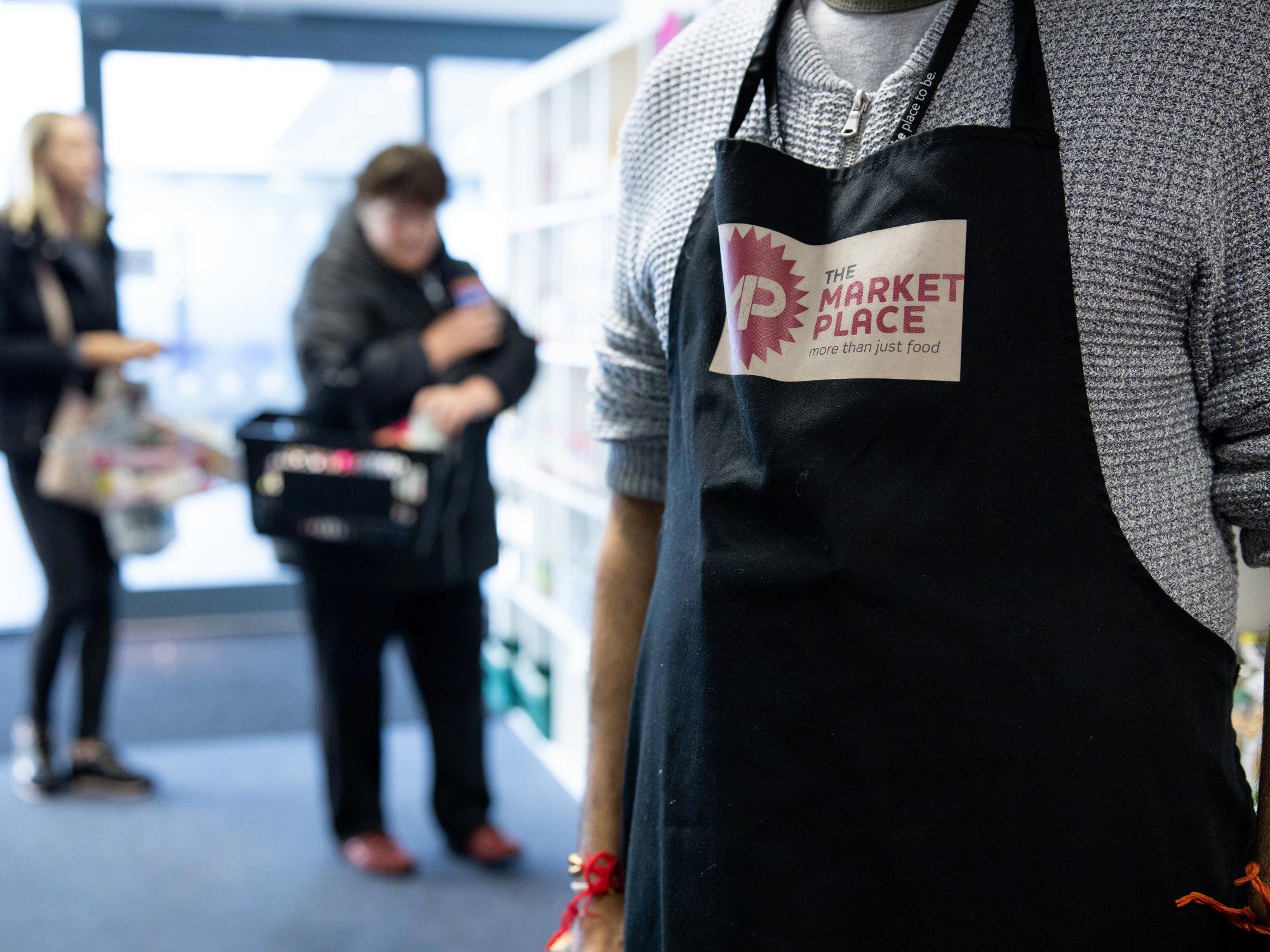 Person wearing an apron with ‘THE MARKET PLACE – more than just food’ logo, standing in a store with shelves and products; two other people are visible in the background, one holding a basket.