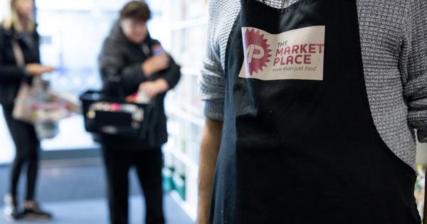 Person wearing an apron with ‘THE MARKET PLACE – more than just food’ logo, standing in a store with shelves and products; two other people are visible in the background, one holding a basket.