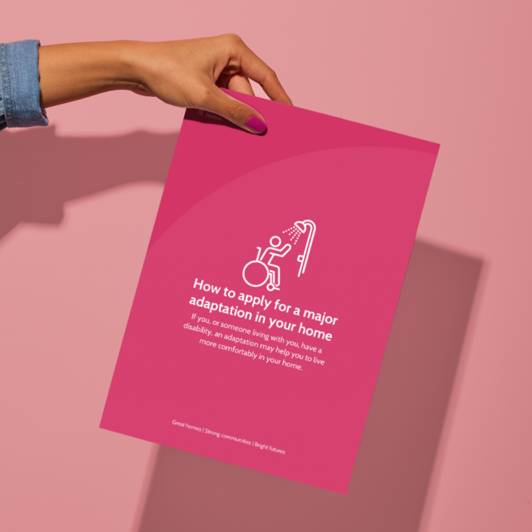 Hand holding a pink flyer against a pink background. The flyer features an icon of a person in a wheelchair under a showerhead and text about applying for a major home adaptation for people with disabilities.