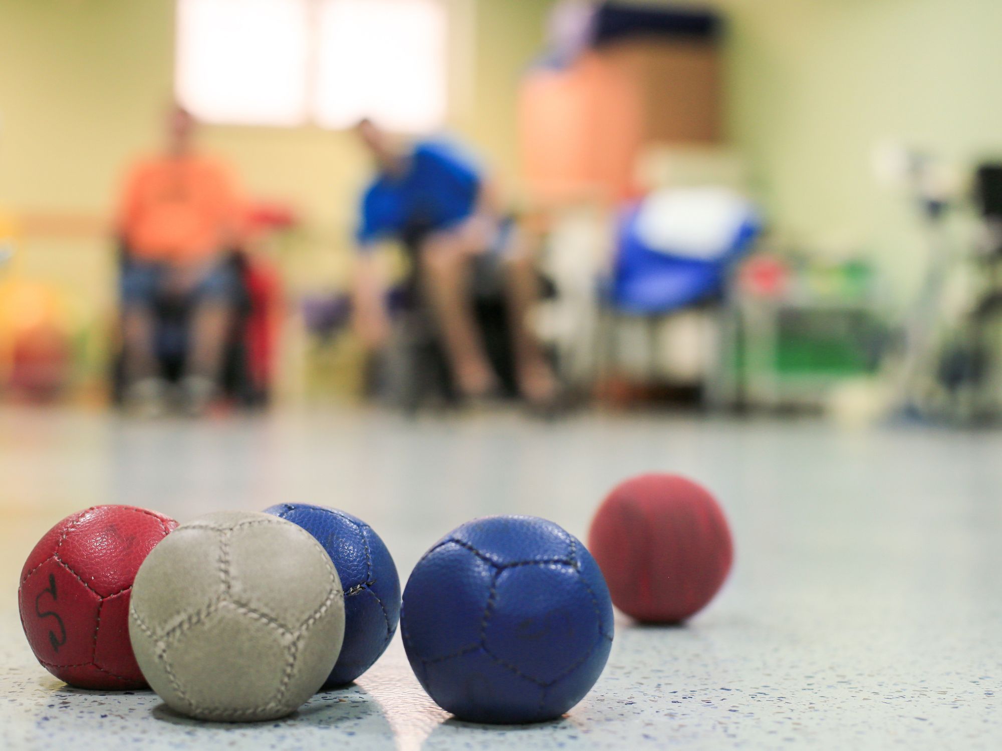 Several boccia balls in red, blue, and white are scattered on a smooth indoor floor in the foreground. The background shows a blurred setting with chairs and two people seated, suggesting an indoor sports or recreational environment. The focus is on the boccia balls, indicating a game or activity in progress.