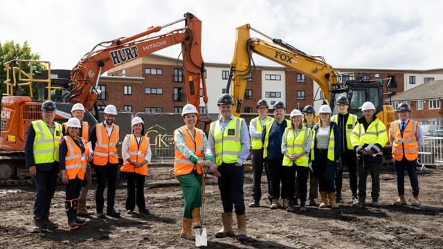 Group of construction workers and officials in high-visibility gear on an urban construction site, with two excavators labelled 'HURT' and 'FOX' in the background.