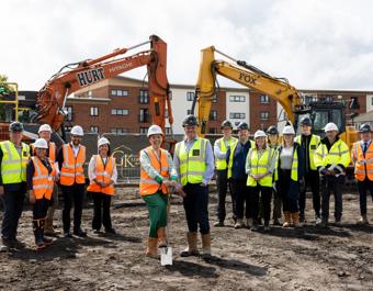 Group of construction workers and officials in high-visibility gear on an urban construction site, with two excavators labelled 'HURT' and 'FOX' in the background.