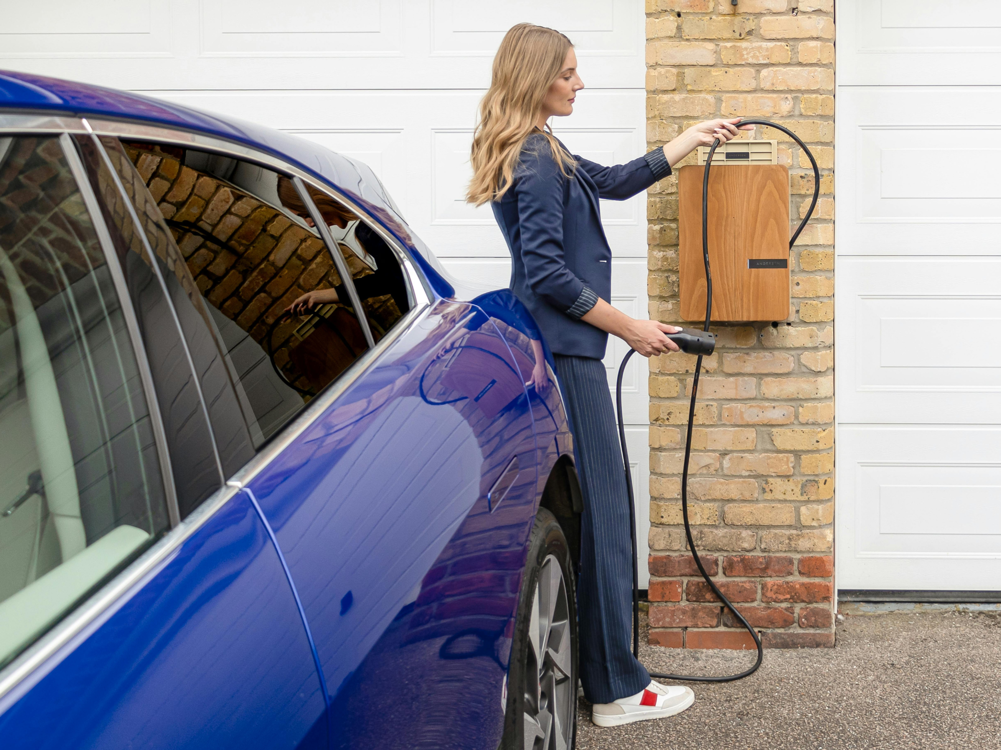 A blue electric car parked on a driveway next to a brick house, with a person connecting a charging cable to a wall-mounted EV charging point.