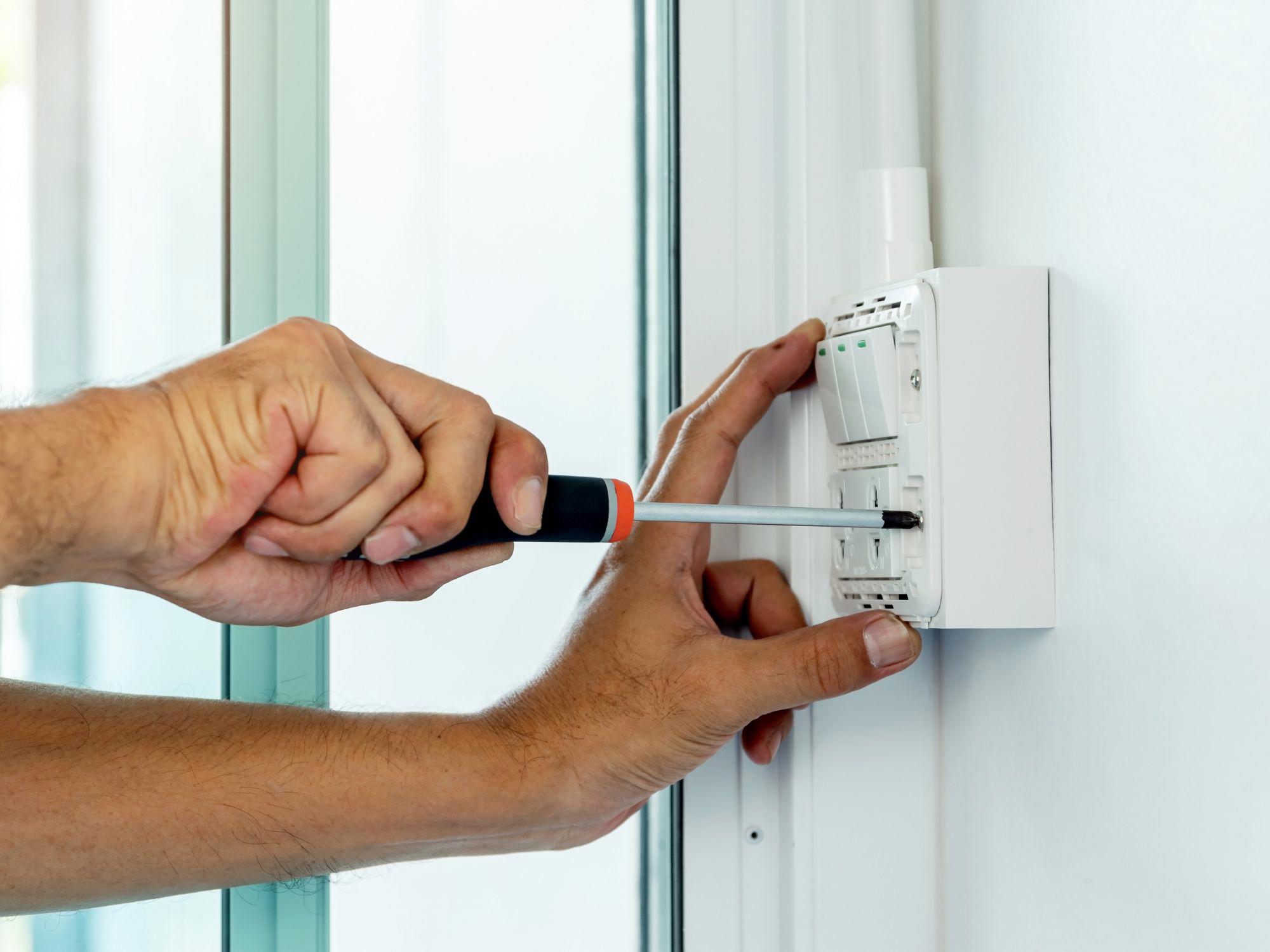 Close-up of hands using a screwdriver to install or repair an electrical wall switch next to a glass door or window.