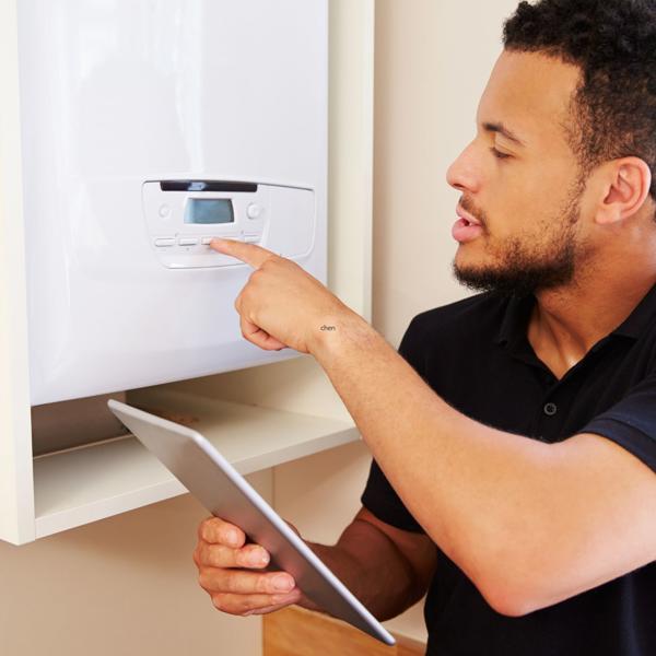 Person using a tablet while pointing at the control panel of a wall-mounted boiler or heating system, possibly for repairs or adjustments.