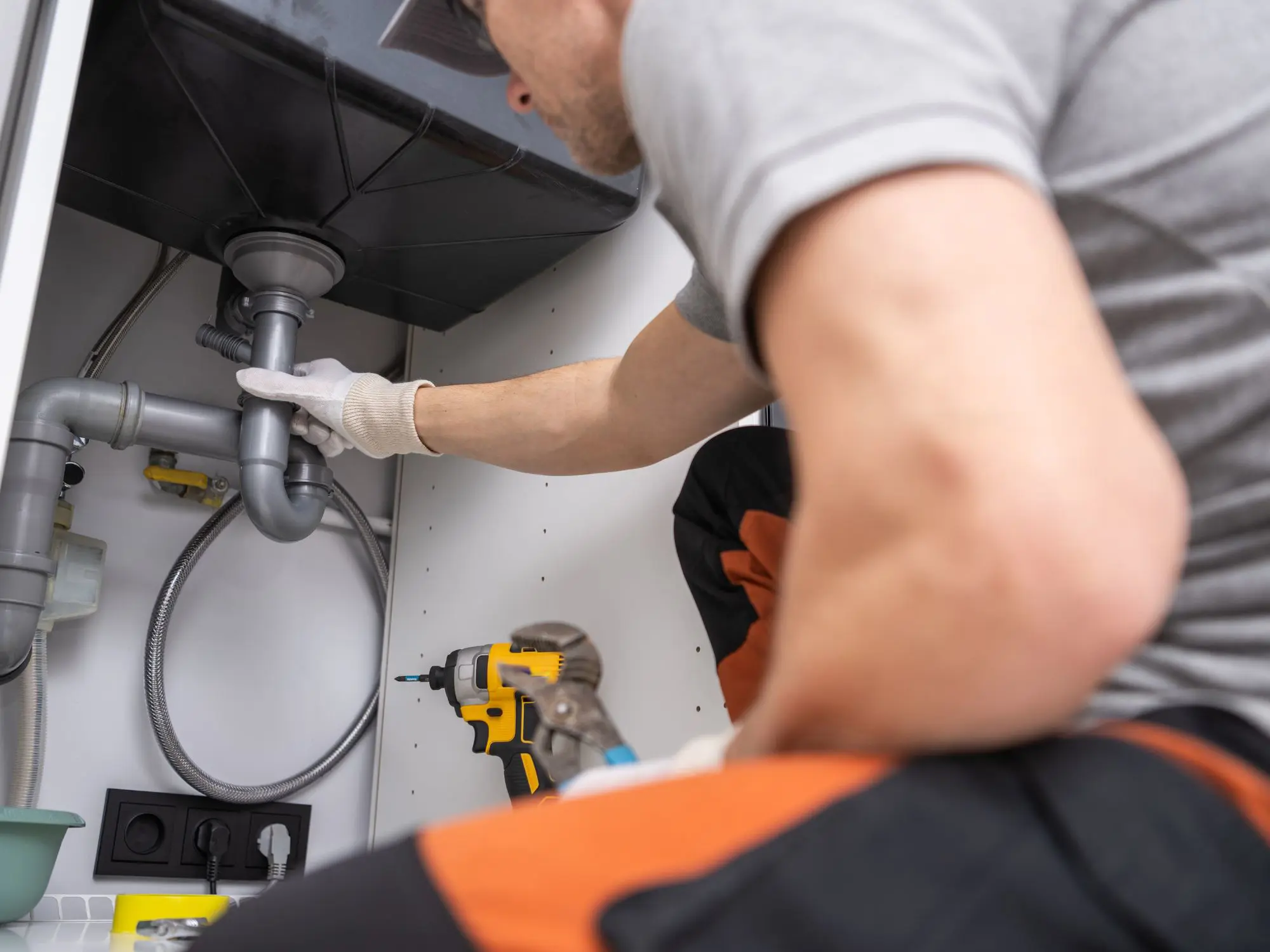 Person working under a sink, pointing at plumbing pipes with tools like a drill and pliers nearby, suggesting plumbing maintenance or repair
