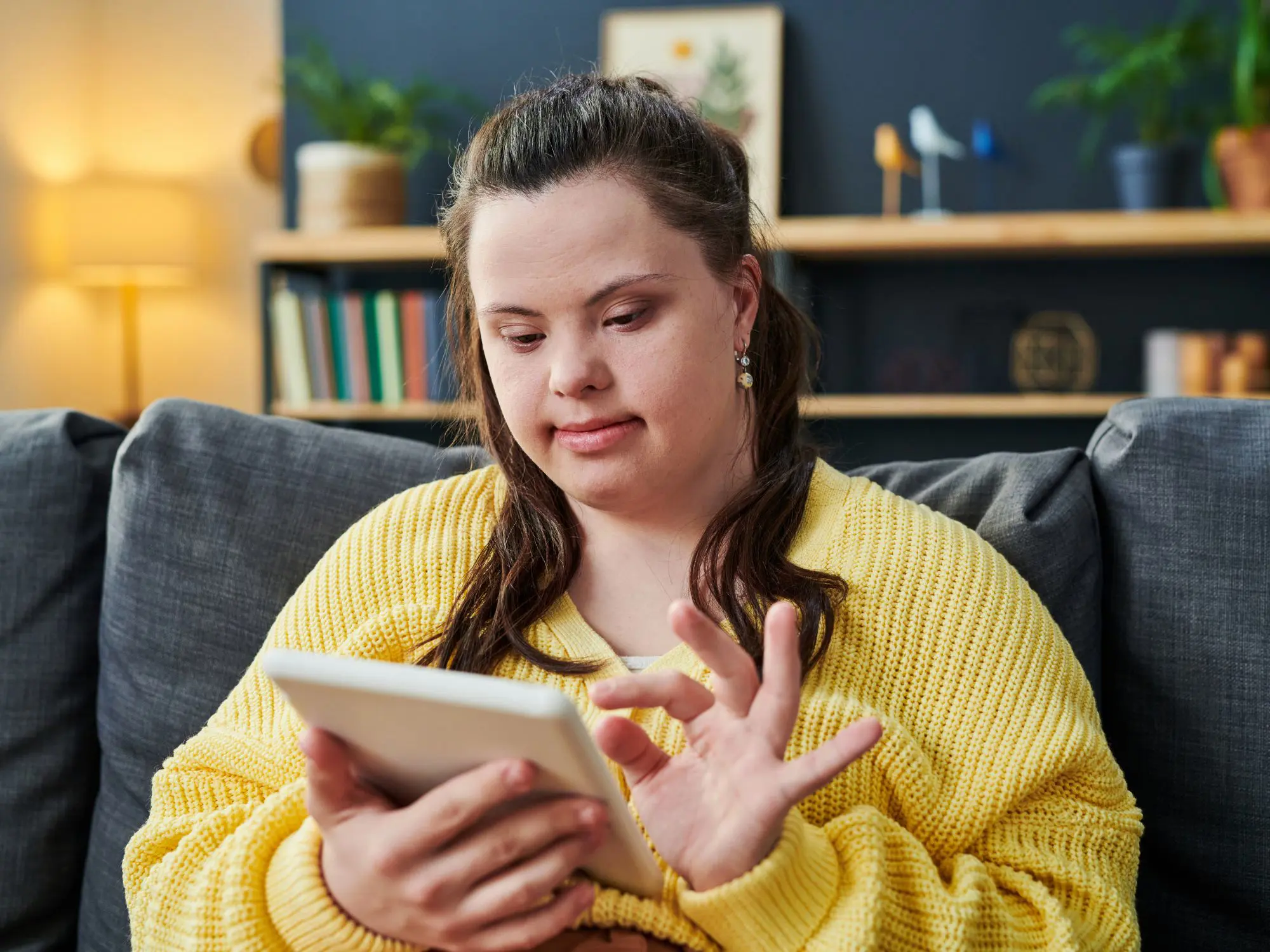 Person in a yellow sweater sitting on a couch and using a tablet. Background includes shelves with books, plants, decorative items, and a lamp providing warm lighting.