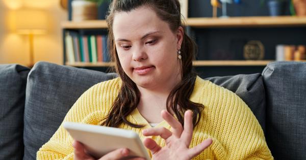 Person in a yellow sweater sitting on a couch and using a tablet. Background includes shelves with books, plants, decorative items, and a lamp providing warm lighting.