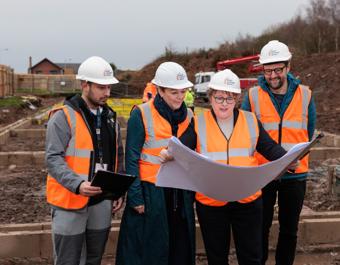 Four people in white hard hats and orange safety vests stand at a construction site, reviewing plans. One holds a large sheet of paper. Behind them are construction materials, partially built structures, and a house in the distance.