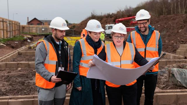 Four people in white hard hats and orange safety vests stand at a construction site, reviewing plans. One holds a large sheet of paper. Behind them are construction materials, partially built structures, and a house in the distance.