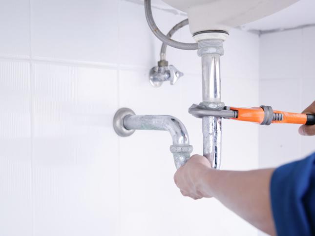 Person using an orange-handled wrench to adjust a metallic pipe connection under a sink, with white tiled walls in the background. Person using an orange-handled wrench to adjust a metallic pipe connection under a sink, with white tiled walls in the background.