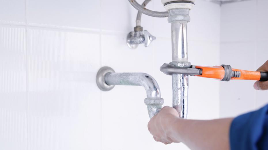 Person using an orange-handled wrench to adjust a metallic pipe connection under a sink, with white tiled walls in the background. Person using an orange-handled wrench to adjust a metallic pipe connection under a sink, with white tiled walls in the background.