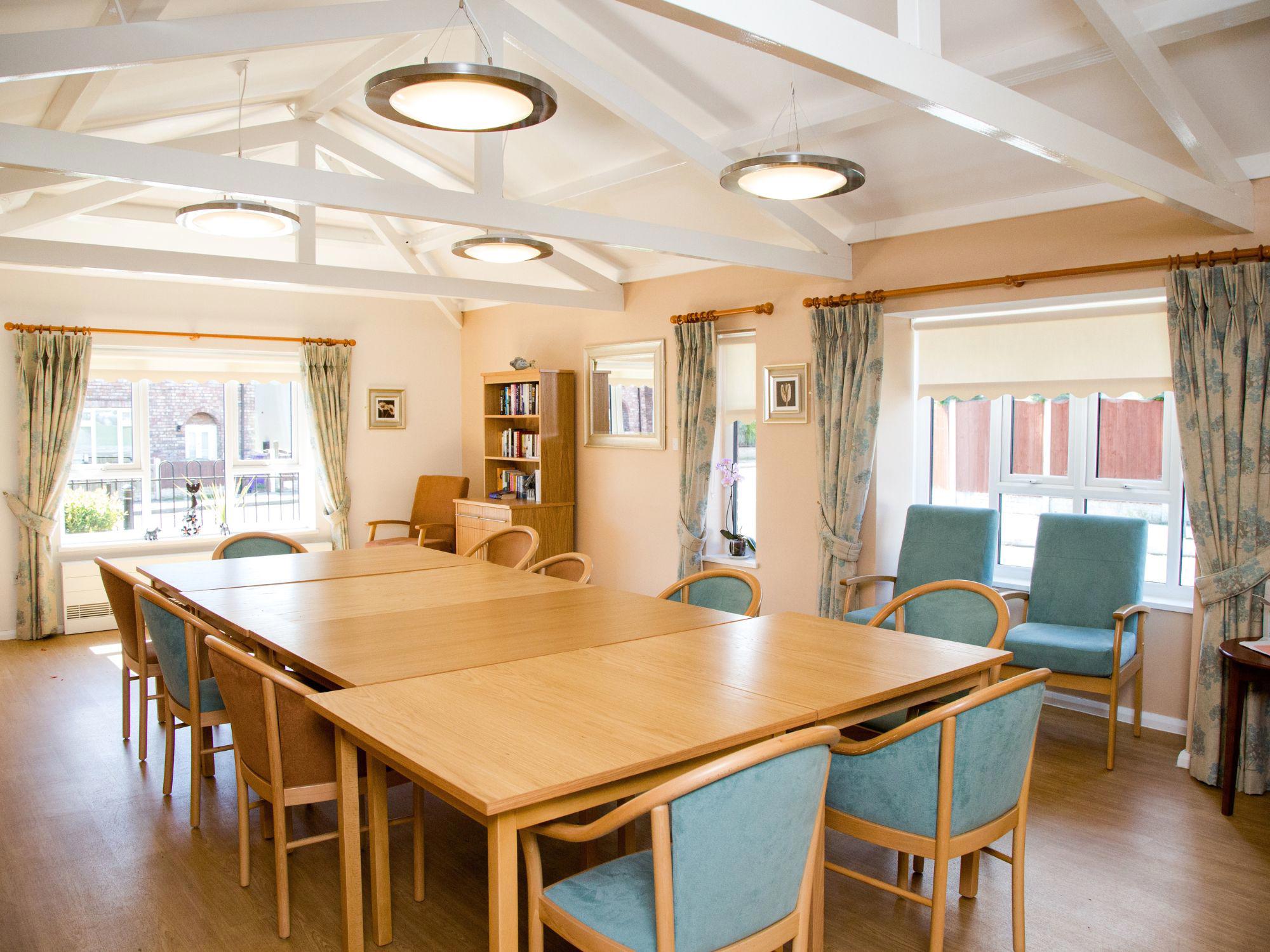 Bright room with high ceilings, exposed white beams, and circular ceiling lights. Two large wooden tables are surrounded by light blue upholstered chairs. Windows with light curtains and a bookshelf filled with books are visible.