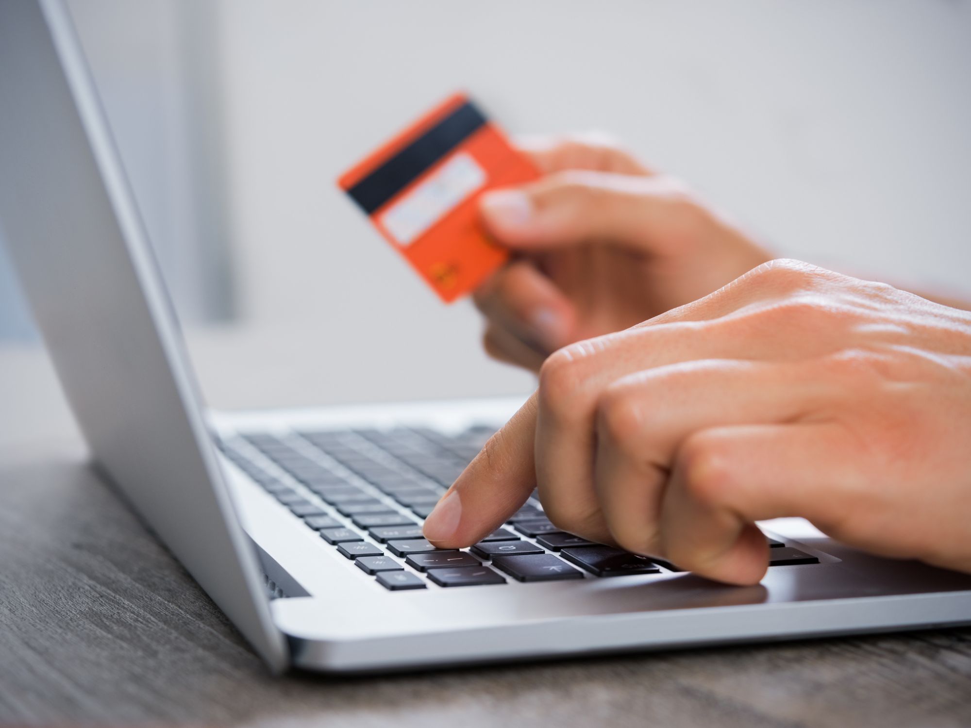 Close-up of hands using a laptop keyboard, with one hand typing and the other holding an orange credit card, representing online payment or banking activity.