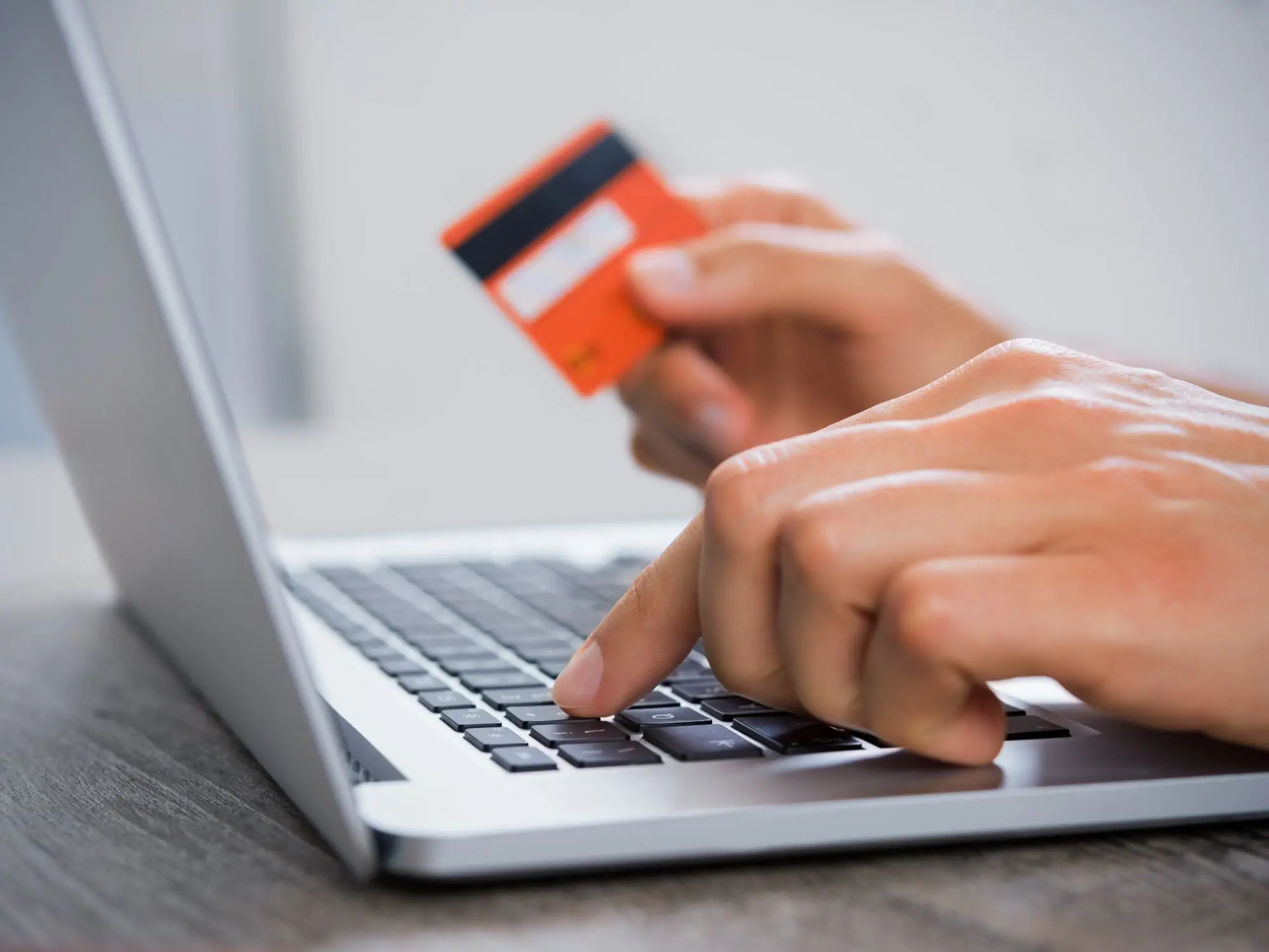 Close-up of hands using a laptop keyboard, with one hand typing and the other holding an orange credit card, representing online payment or banking activity.