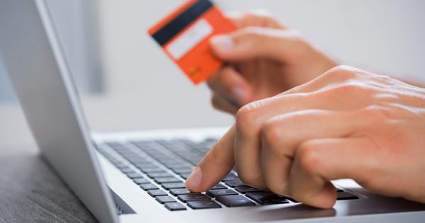 Close-up of hands using a laptop keyboard, with one hand typing and the other holding an orange credit card, representing online payment or banking activity.