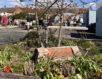 A community garden at Grow Speke with spring flowers and plants in the foreground and a small tree planted in a raised bed. A wooden sign at the front reads “Grow Speke.” In the background, people are gathered around tables and activities on a sunny day, with houses and garden structures visible behind them.