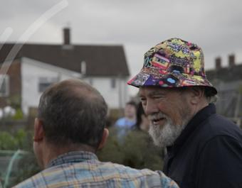 wo people interacting in an outdoor community setting. One wears a plaid shirt and faces away from the camera, while the other, facing the camera, wears a colorful patterned hat. Houses and other people are visible in the background under an overcast sky.