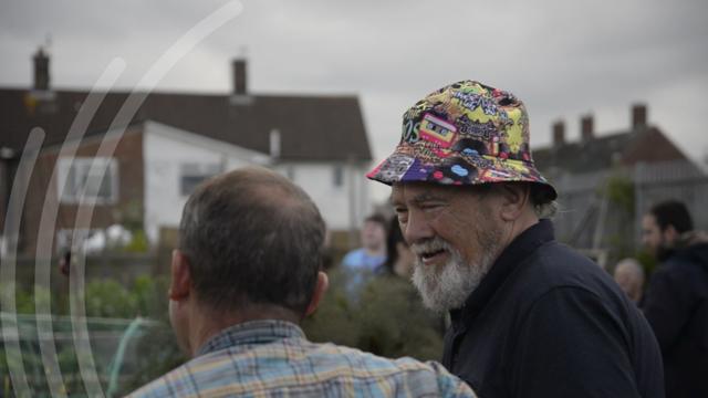 wo people interacting in an outdoor community setting. One wears a plaid shirt and faces away from the camera, while the other, facing the camera, wears a colorful patterned hat. Houses and other people are visible in the background under an overcast sky.