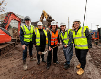 Five people in high-visibility vests and hard hats standing on a muddy construction site, with one person holding a shovel. Construction vehicles, including an excavator and a red Kubota digger, are visible in the background, indicating a groundbreaking event.