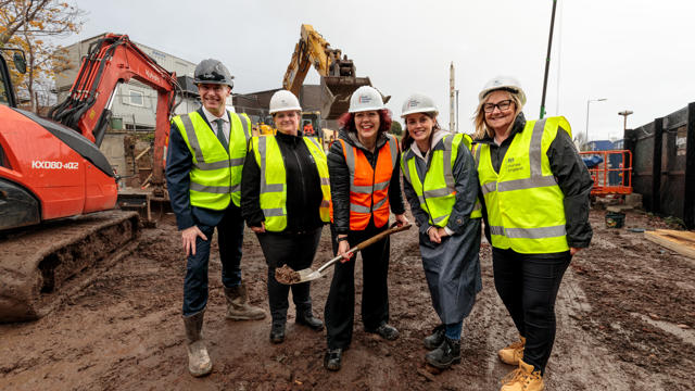 Five people in high-visibility vests and hard hats standing on a muddy construction site, with one person holding a shovel. Construction vehicles, including an excavator and a red Kubota digger, are visible in the background, indicating a groundbreaking event.