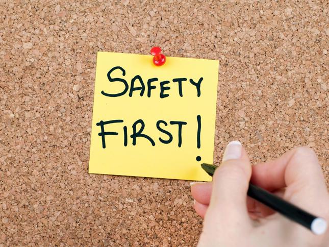 Yellow sticky note with 'SAFETY FIRST!' written in black marker pinned to a corkboard with a red pushpin, and a hand holding a black pen nearby. Yellow sticky note with 'SAFETY FIRST!' written in black marker pinned to a corkboard with a red pushpin, and a hand holding a black pen nearby.