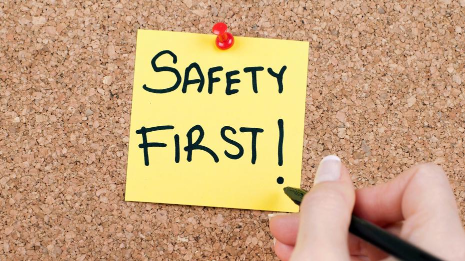 Yellow sticky note with 'SAFETY FIRST!' written in black marker pinned to a corkboard with a red pushpin, and a hand holding a black pen nearby. Yellow sticky note with 'SAFETY FIRST!' written in black marker pinned to a corkboard with a red pushpin, and a hand holding a black pen nearby.