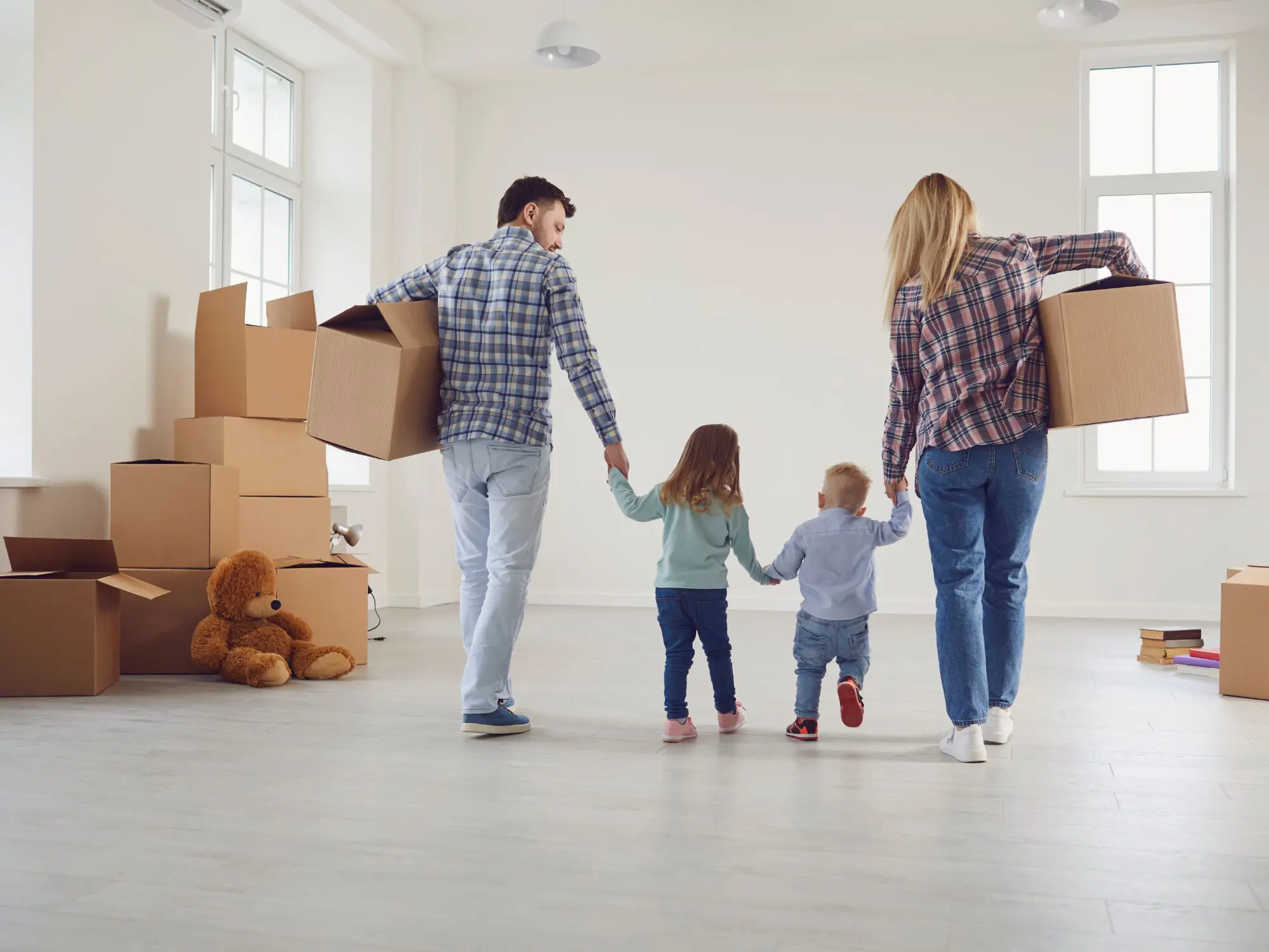 A family of four—two adults and two children—in a bright, empty room with large windows and several cardboard boxes; the adults are holding the children’s hands and carrying boxes, with a large teddy bear on the floor beside stacked boxes.