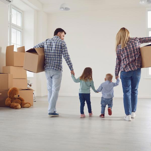 A family of four—two adults and two children—in a bright, empty room with large windows and several cardboard boxes; the adults are holding the children’s hands and carrying boxes, with a large teddy bear on the floor beside stacked boxes.