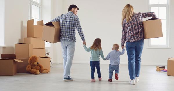 A family of four—two adults and two children—in a bright, empty room with large windows and several cardboard boxes; the adults are holding the children’s hands and carrying boxes, with a large teddy bear on the floor beside stacked boxes.