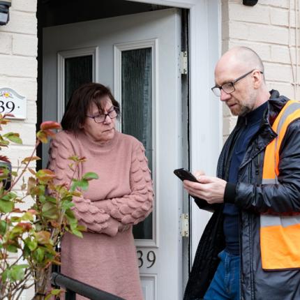 Two individuals standing outside house number 39. One wears a pink sweater and glasses with arms crossed, while the other wears an orange safety vest and shows something on a smartphone.