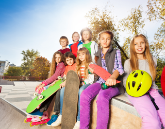 A group of young people sitting on the edge of a skatepark ramp, holding skateboards and a bright yellow helmet, with trees and blue sky in the background.