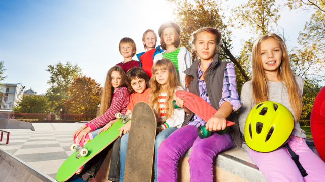 A group of young people sitting on the edge of a skatepark ramp, holding skateboards and a bright yellow helmet, with trees and blue sky in the background.